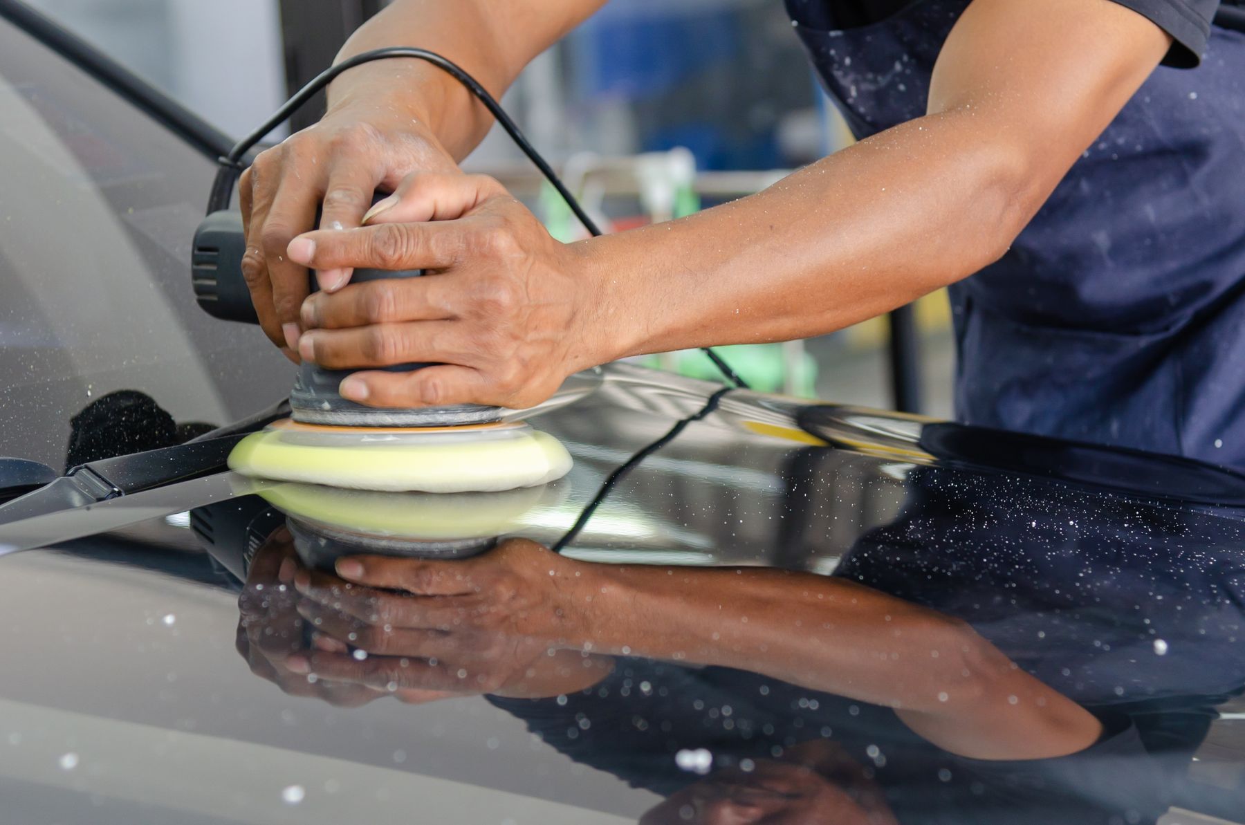 A detailer's hands guiding a polisher across a car's dark hood.